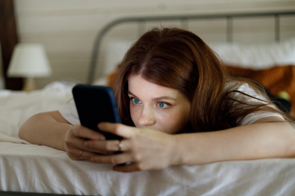 ragazza con gli occhi chiari stesa sul letto che guarda il cellulare